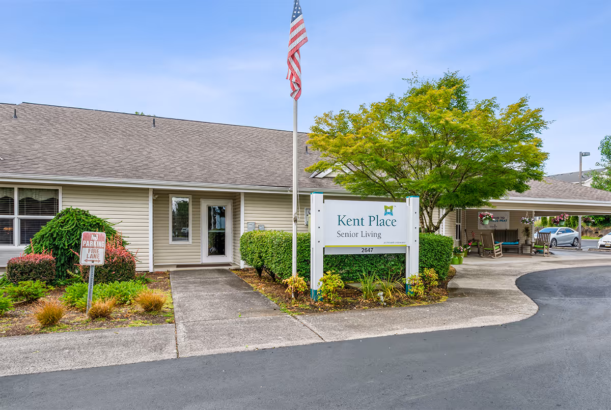 Exterior view of Kent Place Senior Living facility showing the entrance with a sidewalk, landscaping with bushes and a tree, an American flag on a flagpole, and a sign displaying the facility name and address. There is a covered area with rocking chairs and hanging flower pots to the right.