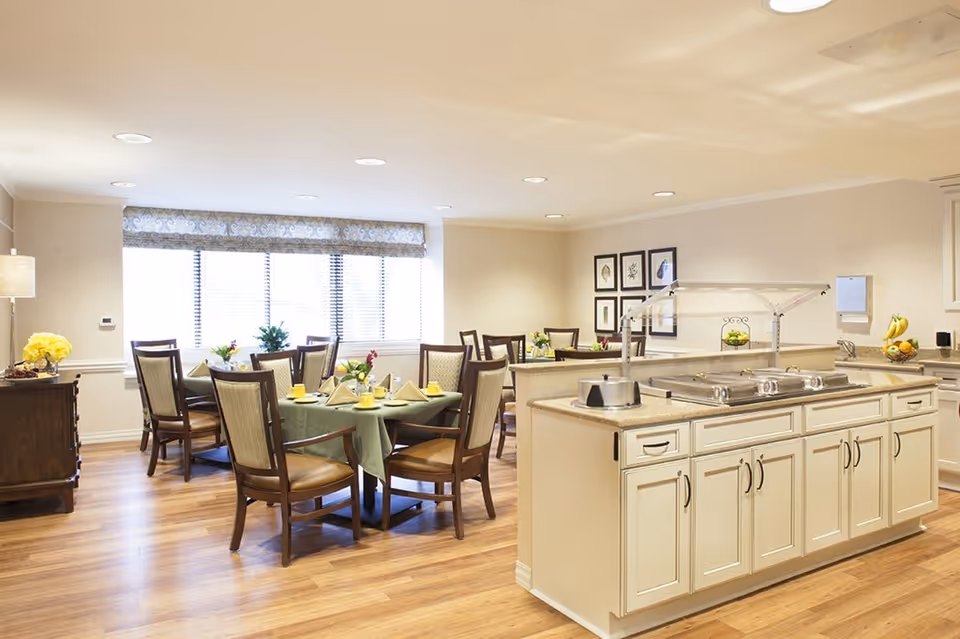 A bright dining room in a senior living facility with several round tables covered with green tablecloths, each set with yellow cups, napkins, and small flower arrangements. Wooden chairs with cushioned seats surround the tables. In the foreground, there is a buffet counter with serving trays and a sneeze guard. The room has wooden flooring, large windows with blinds, and framed artwork on the walls.
