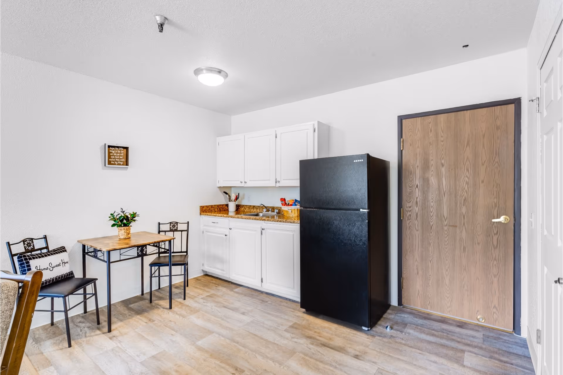 A small kitchen area with white cabinets, a black refrigerator, and a wooden door. There is a small dining table with two chairs, one with a 'Home Sweet Home' pillow, and a small plant on the table. The floor is light wood, and the walls are white with a small framed quote hanging.