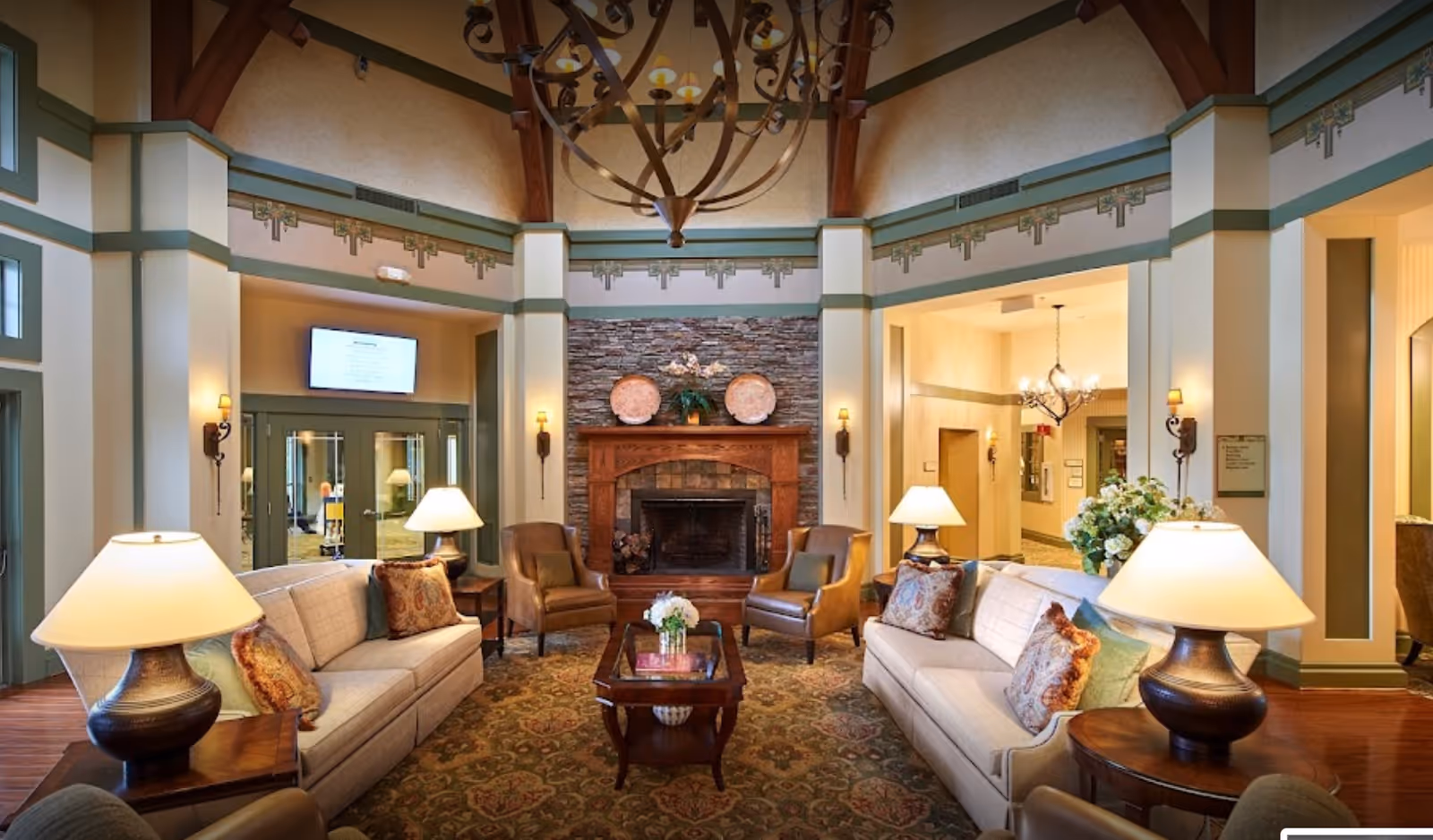A spacious and warmly decorated living room area with two beige sofas adorned with patterned cushions, two brown leather armchairs, a wooden coffee table with a flower arrangement, and a stone fireplace with decorative plates and a plant on the mantel. The room features high ceilings with exposed wooden beams, elegant wall sconces, and multiple table lamps providing soft lighting.