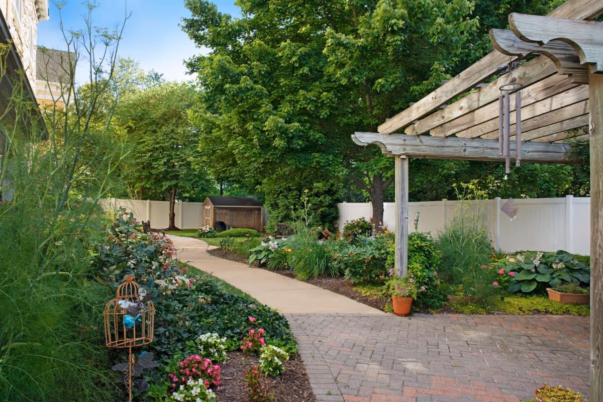 A peaceful outdoor garden area with a paved walkway winding through lush greenery and colorful flowers. There is a wooden pergola on the right side with a wind chime hanging from it, and a small wooden shed is visible in the background. The area is enclosed by a white fence and shaded by large trees.