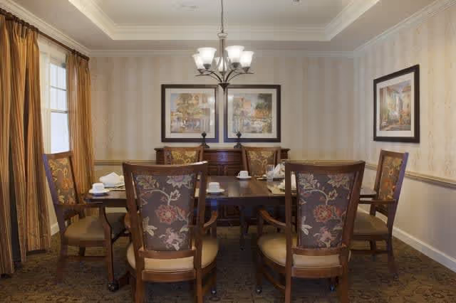 A dining room with a rectangular wooden table surrounded by six upholstered chairs with floral patterns. The table is set with white cups and napkins. The room has beige patterned wallpaper, two framed paintings on the far wall, a chandelier hanging above the table, and windows with brown curtains on the left side.