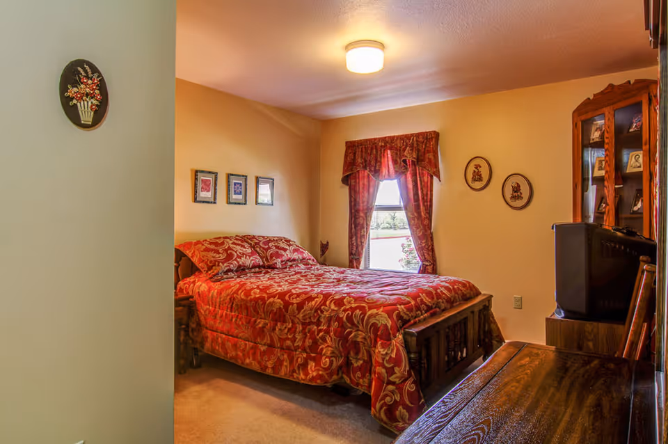 A cozy bedroom featuring a wooden bed with red patterned bedding, a window with curtains, and wooden furniture.