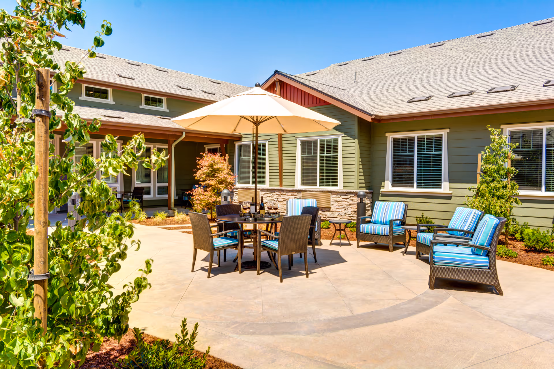 Outdoor patio area at The Vineyard at Fountaingrove Memory Care with a round table and four chairs under a large beige umbrella, surrounded by additional seating with blue striped cushions. The patio is adjacent to a green building with multiple windows and landscaping including small trees and shrubs.