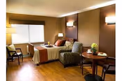 A cozy bedroom in Baldwin Health Center featuring a single bed with white and brown bedding, a cushioned armchair, a small round table with two chairs, a lamp, and a window with blinds. The room has wooden flooring and warm brown walls with soft lighting fixtures.