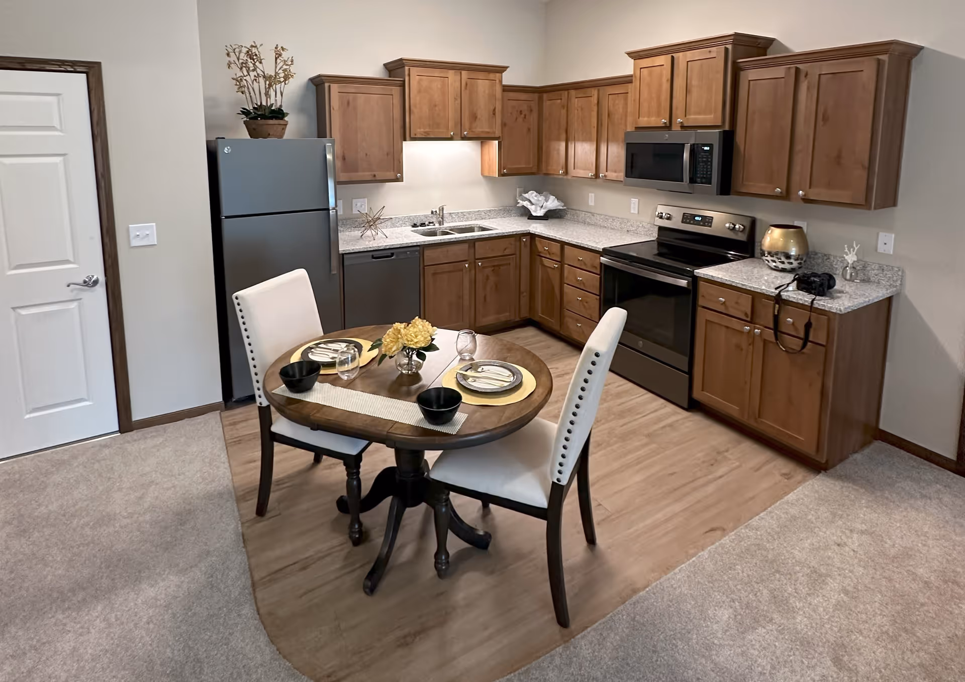 A compact kitchen area with wooden cabinets, a stainless steel refrigerator, dishwasher, stove, and microwave. In front of the kitchen is a small round wooden dining table set for two with white cushioned chairs, yellow placemats, plates, bowls, and glasses. The floor transitions from carpet to wood in the kitchen area. Decorative items are placed on the countertops and above the refrigerator.