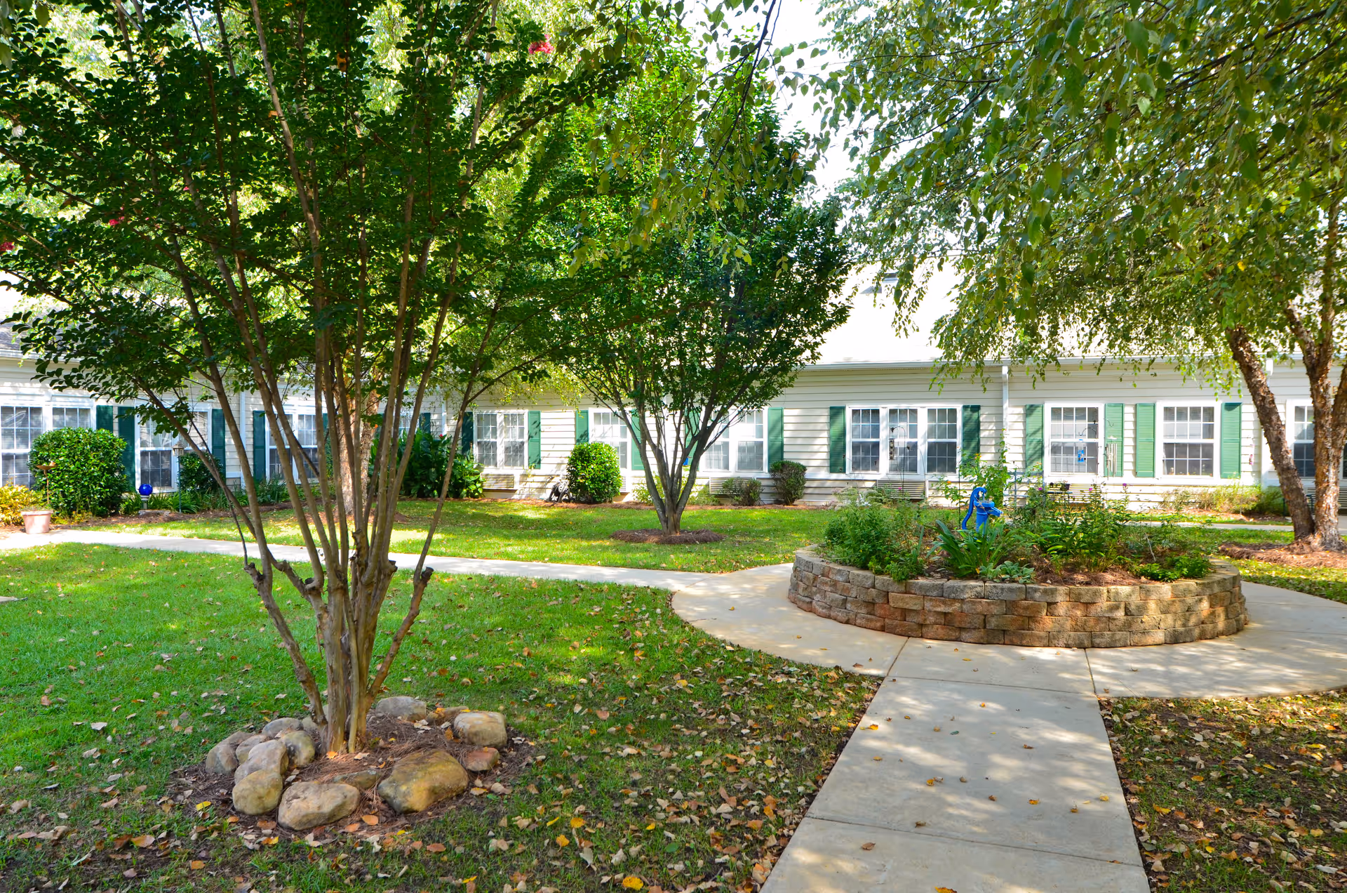 A peaceful outdoor garden area at Morningside of Anderson featuring a circular stone planter with greenery and a small blue sculpture in the center. Surrounding the planter are concrete walkways and well-maintained grass with trees providing shade. In the background, there is a single-story building with white siding and green shutters on the windows.