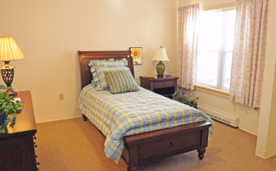 A cozy bedroom with a single wooden bed featuring a plaid bedspread and multiple pillows. There is a wooden nightstand with a green lamp and a framed sunflower picture on the wall. A window with patterned curtains allows natural light into the room. A dresser with a lamp and a plant is partially visible on the left side.