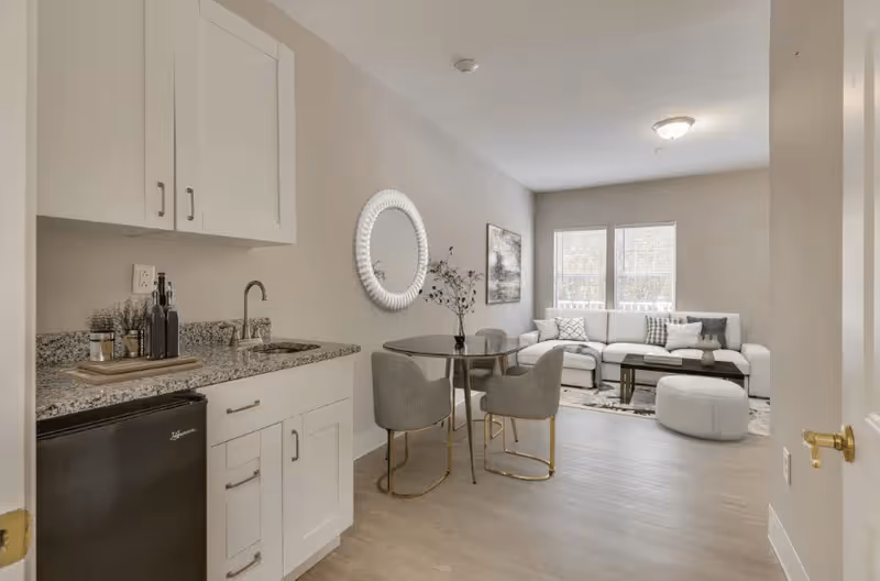 A modern living space featuring a small kitchenette with white cabinets, granite countertop, and a mini refrigerator on the left. Adjacent to the kitchenette is a round dining table with four gray upholstered chairs. In the background, there is a bright living room area with a large white sectional sofa, a coffee table, a round ottoman, and two large windows letting in natural light. The walls are painted a light beige color, and there is a decorative round mirror and a framed artwork on the wall.