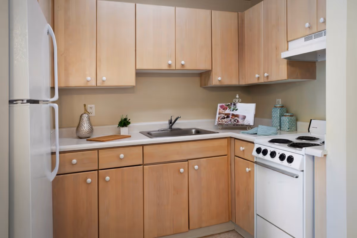 A compact kitchen with light wood cabinets, a white refrigerator on the left, a stainless steel sink in the center, and a white electric stove with four burners on the right. The countertop has a decorative vase, a small plant, a wooden cutting board, a cookbook on a stand, and three matching canisters. The walls are painted beige.