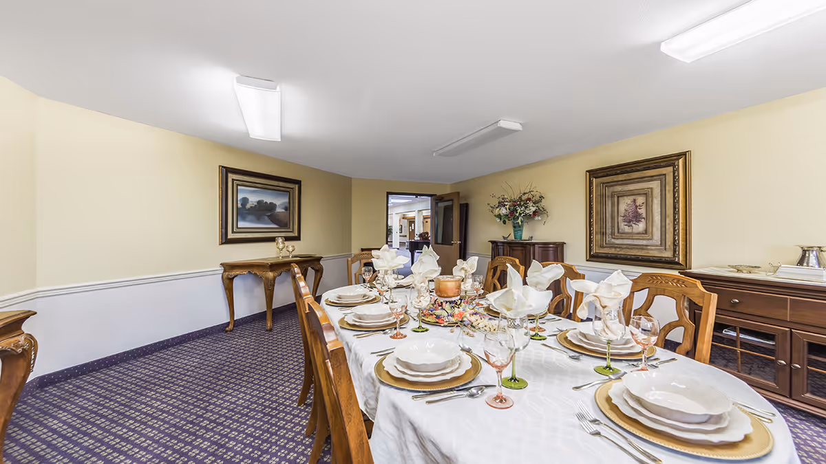 Formal dining room with a long table set with plates, glassware, and folded napkins in a senior living facility.