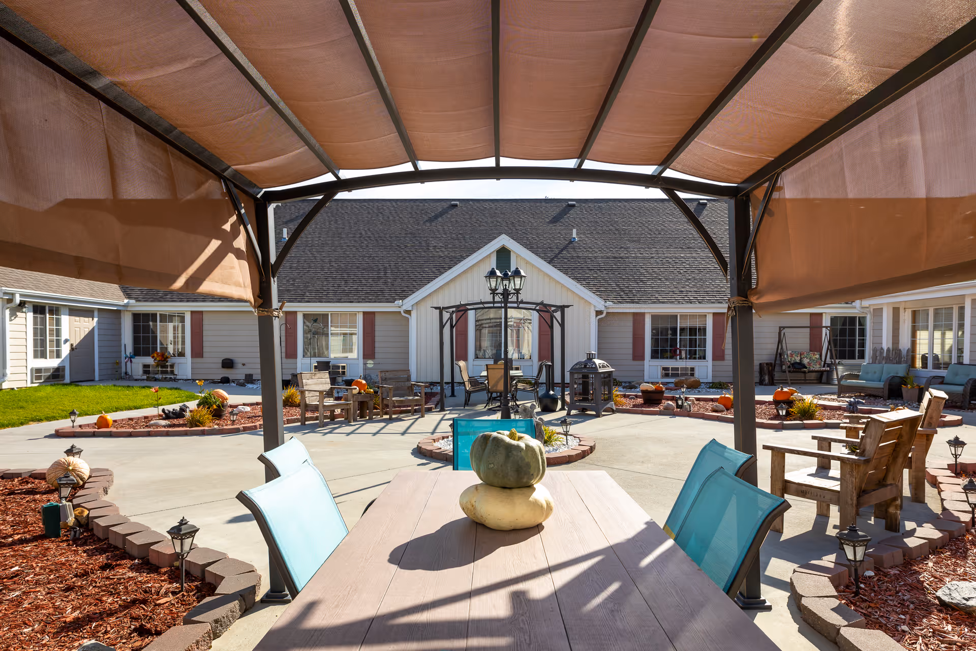 Shaded patio table and chairs under a pergola overlooking a landscaped courtyard with seating, pumpkins, and surrounding single-story facility buildings.