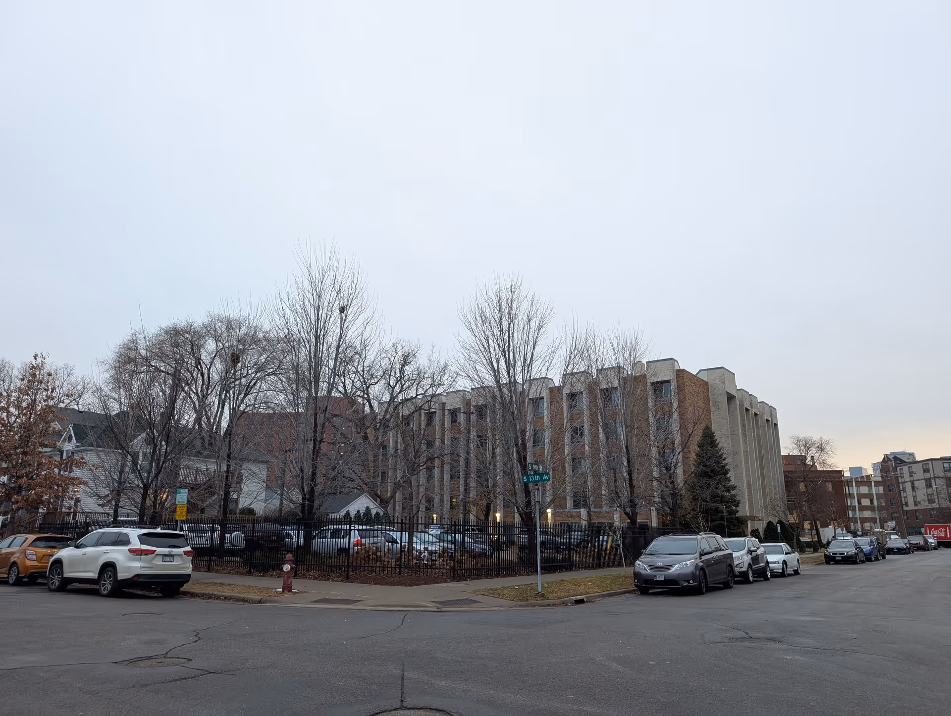 Street view of a multi-story brick and concrete building with several parked cars along the street and a fenced parking lot. Leafless trees are visible around the building under an overcast sky.