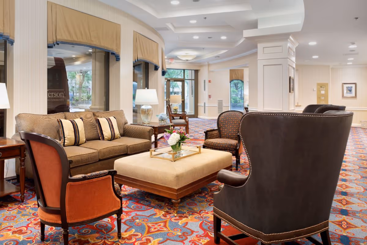 A bright senior living facility lounge with upholstered sofas and chairs arranged around a central ottoman on a colorful patterned carpet.