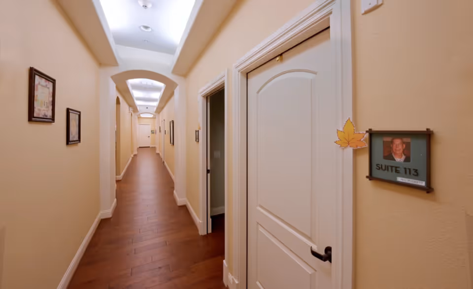 Long interior hallway with wood floors, recessed ceiling lights, framed artwork on the walls, and a door marked "Suite 113" with a resident photo.