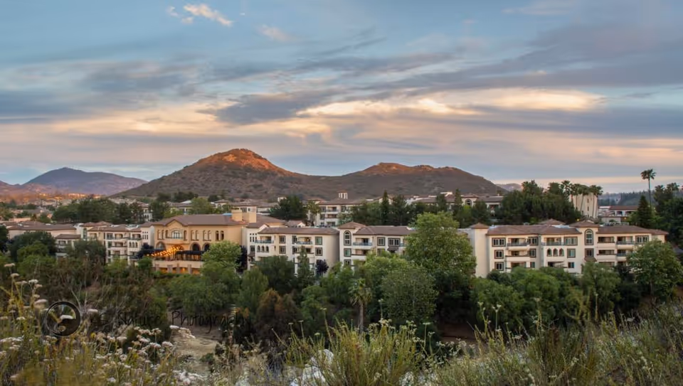 Panoramic exterior view of the Casa de las Campanas senior living campus with multi-story buildings, trees, and hills at sunset.