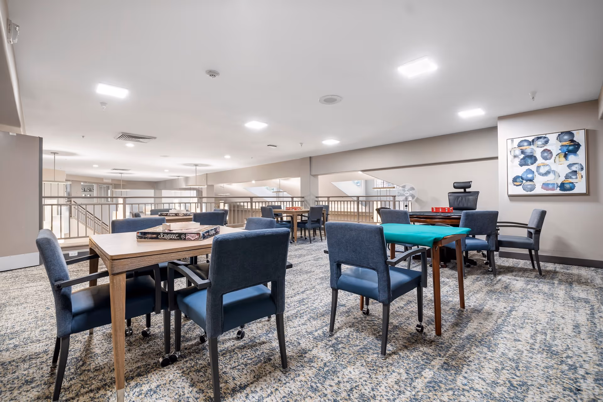 A spacious activity room with several tables and blue upholstered chairs on a patterned carpet. Some tables have board games like Scrabble and card games set up. The room has neutral-colored walls, bright ceiling lights, and a modern abstract painting on one wall. There is a railing along one side overlooking a lower level.