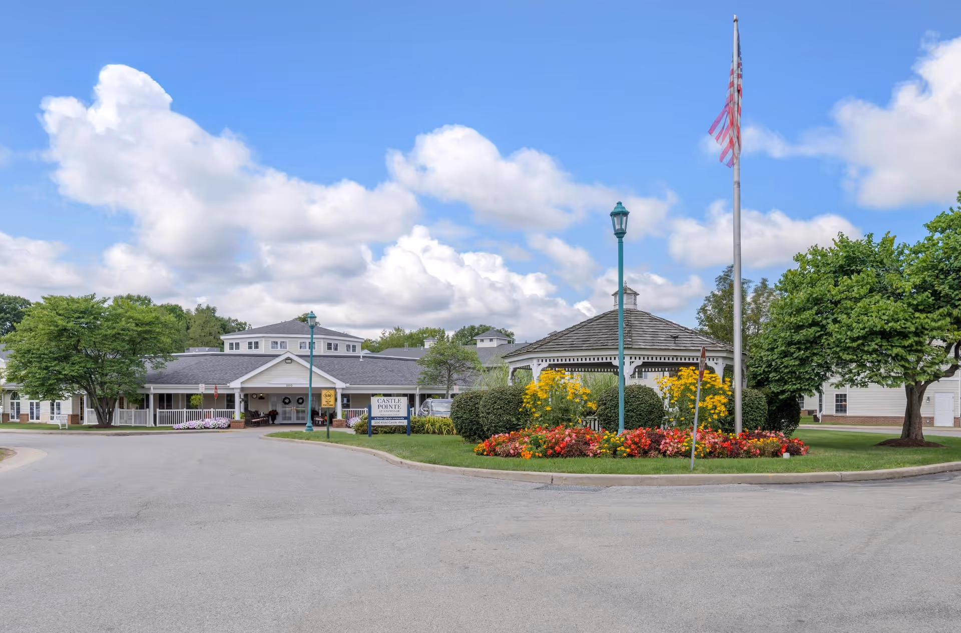 Front exterior view of Castle Pointe at Baywinde senior living facility with a circular driveway, a gazebo surrounded by colorful flowers, an American flag on a flagpole, and trees under a partly cloudy blue sky.