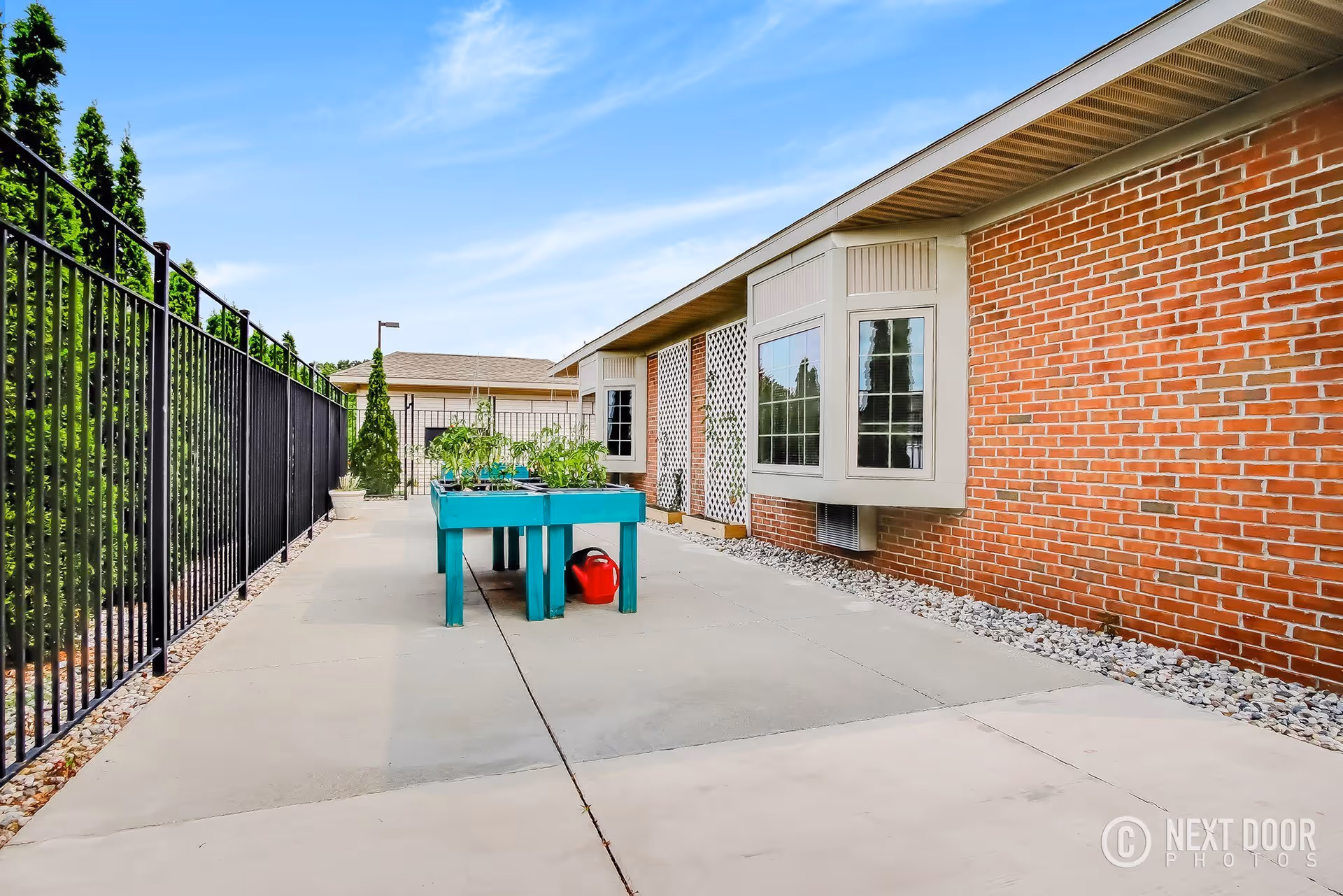 Outdoor patio area at Grandhaven Living Center with a raised garden bed containing plants and a red watering can underneath. The patio is paved with concrete and bordered by a black metal fence on one side and a brick building wall with windows on the other. The sky is clear and blue.