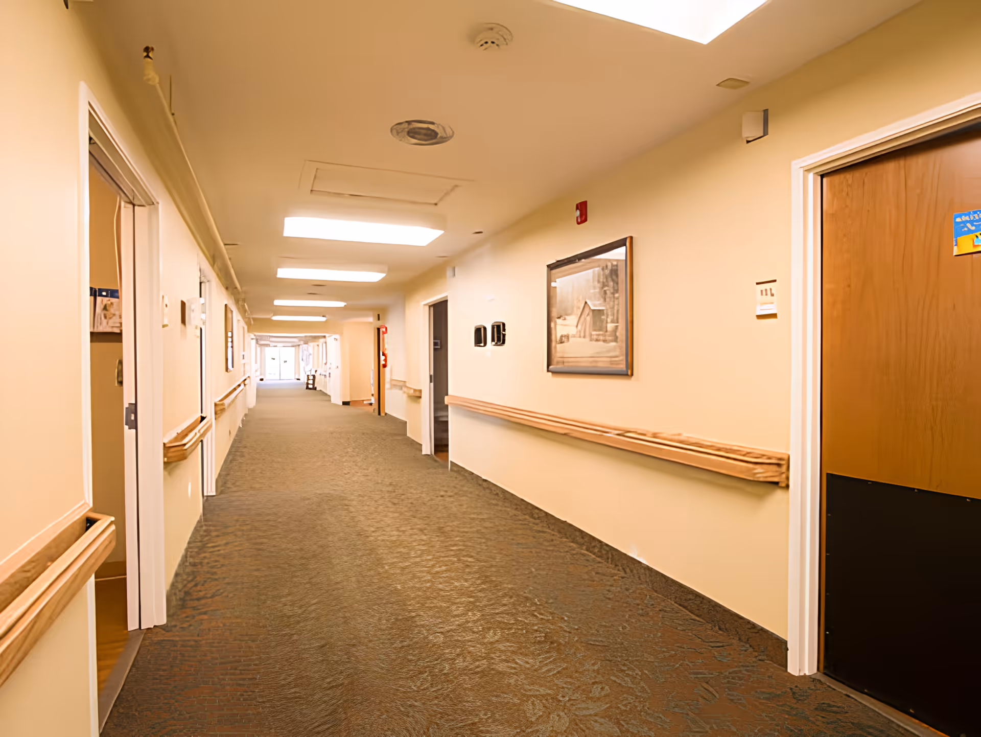 A long, well-lit hallway in a senior living facility with beige walls, carpeted floor, wooden handrails on both sides, several doorways, and a framed picture on the right wall.
