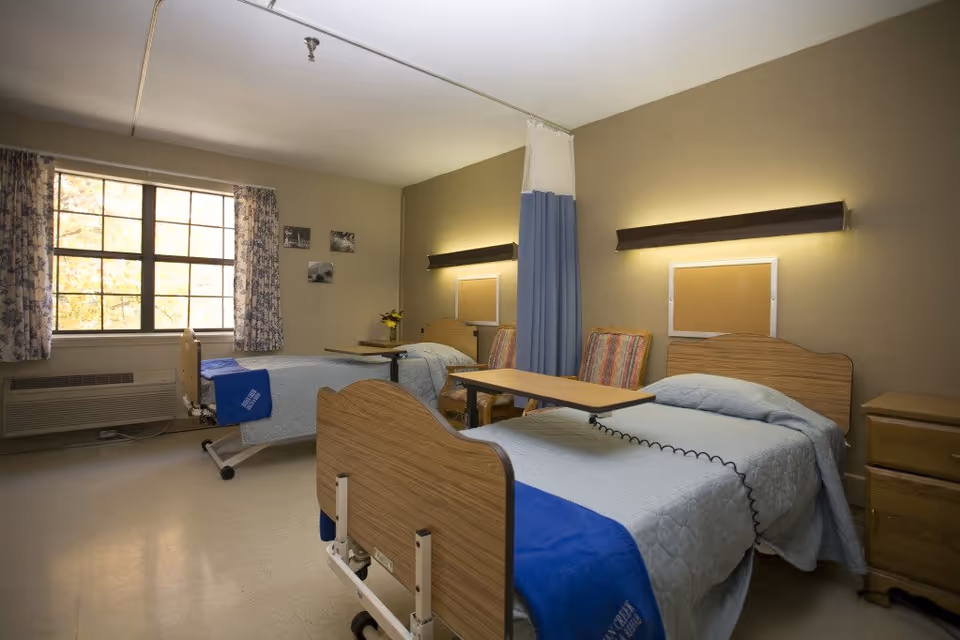 A shared senior living bedroom with two hospital-style beds, overbed tables, chairs, and a window with floral curtains.
