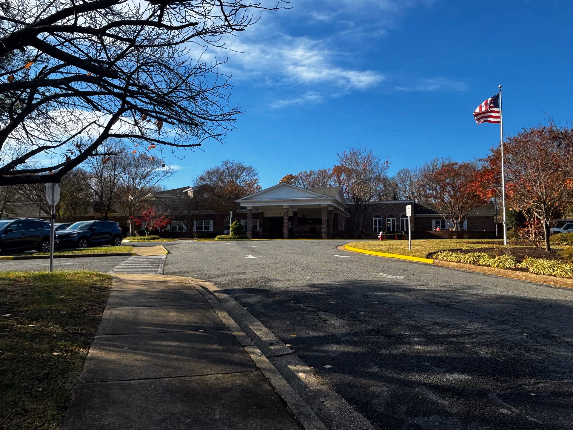 Entrance to a single-story brick retirement community with a circular driveway, American flag, parked cars and trees under a blue sky.