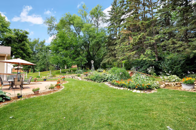 A well-maintained outdoor garden area with a lush green lawn, flower beds bordered by rocks, various plants and flowers, tall trees in the background, and a patio area with a table, chairs, and an umbrella on the left side.