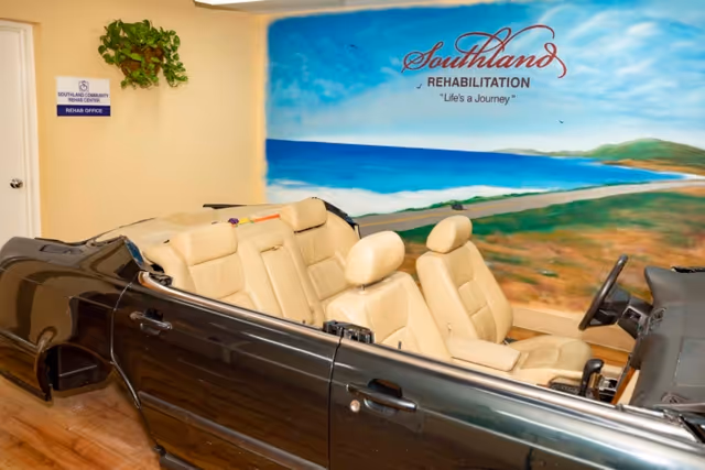 Indoor rehabilitation room featuring a cutaway black car with beige seats used for therapy, set against a large seaside mural reading "Southland Rehabilitation - Life's a Journey."