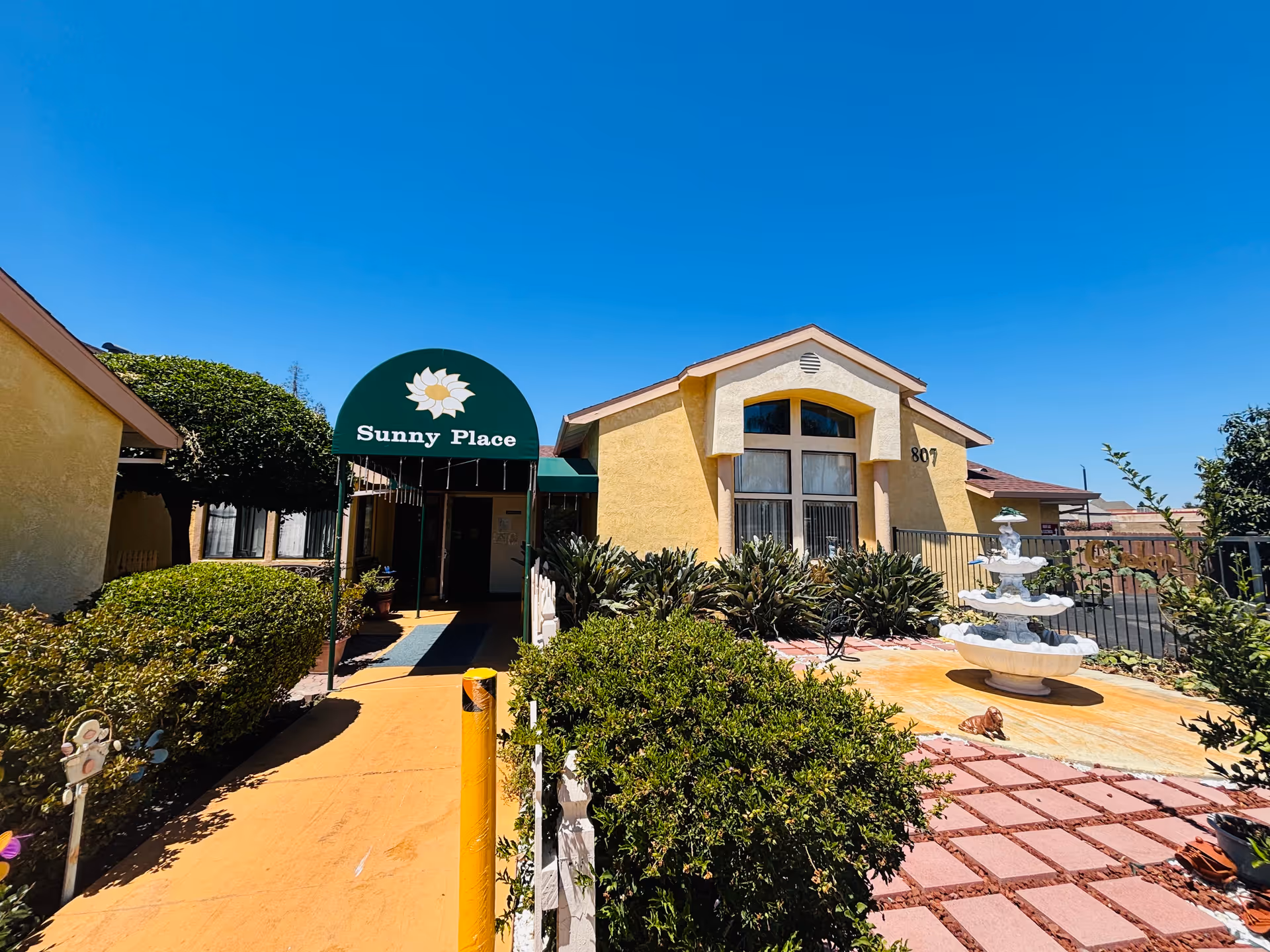 Exterior view of Sunny Place of Stockton, showing a yellow building with a green awning entrance labeled 'Sunny Place'. There are bushes and plants along the walkway leading to the entrance, a white multi-tiered fountain on the right, and a clear blue sky overhead.