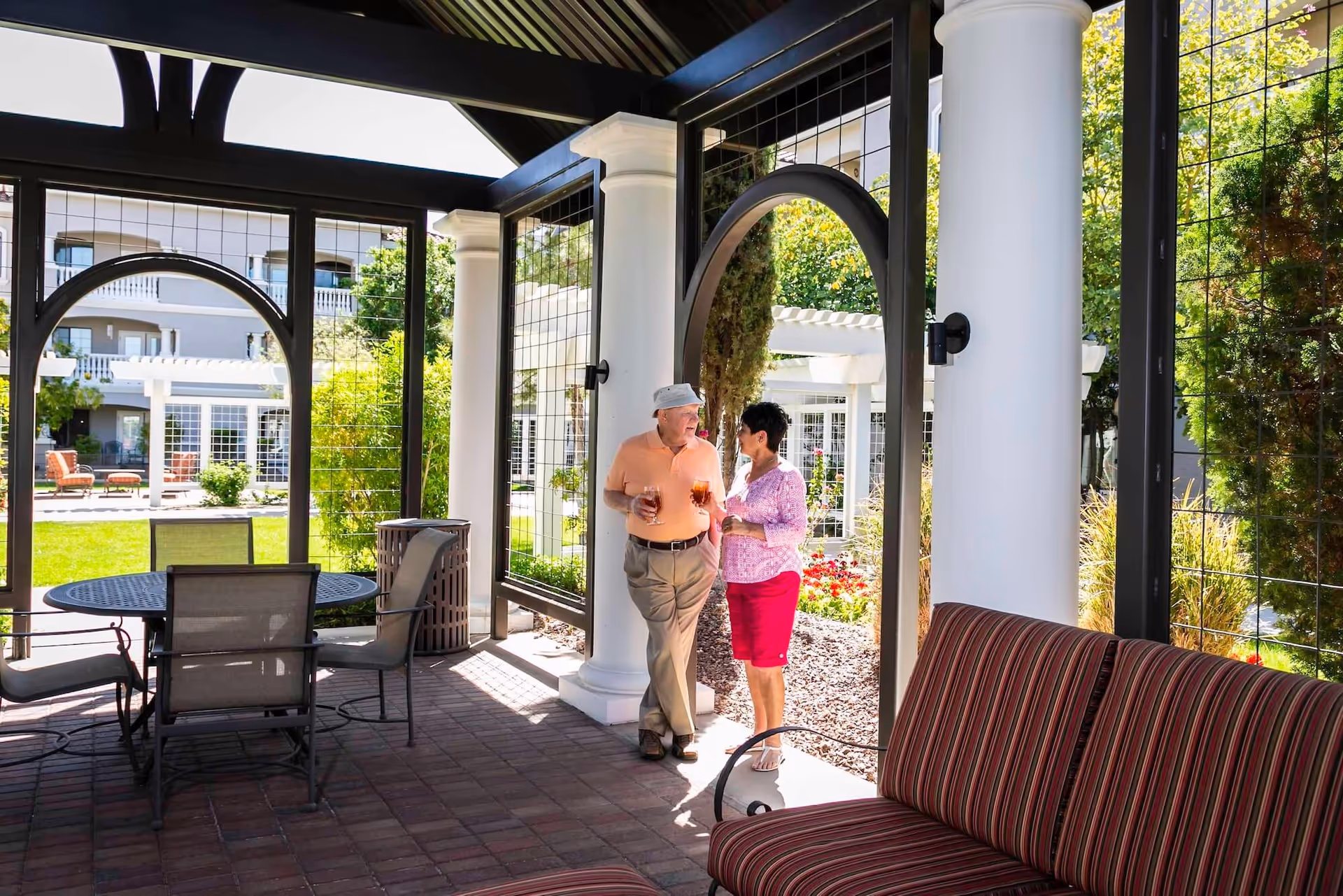 An elderly couple standing and holding drinks under a shaded outdoor patio area with white columns and black metal framing. There is a round table with chairs and a striped cushioned bench nearby. In the background, there is a well-maintained garden and a multi-story building with balconies.
