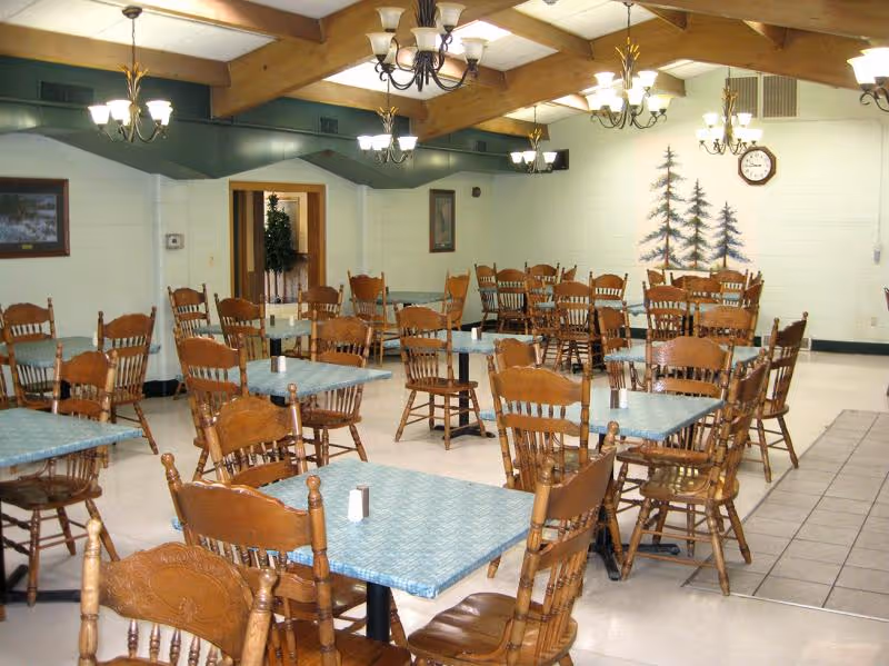 Large dining room with multiple wooden tables and spindle-back chairs under chandeliers and exposed ceiling beams.