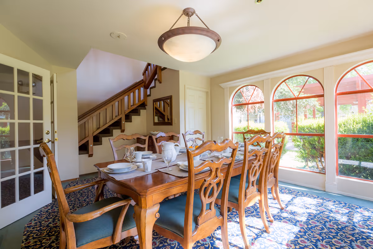 A bright dining room with a wooden dining table set for six, featuring plates, glasses, and cups. The room has large arched windows letting in natural light and offering a view of greenery outside. A wooden staircase and a glass-paneled door are visible in the background.