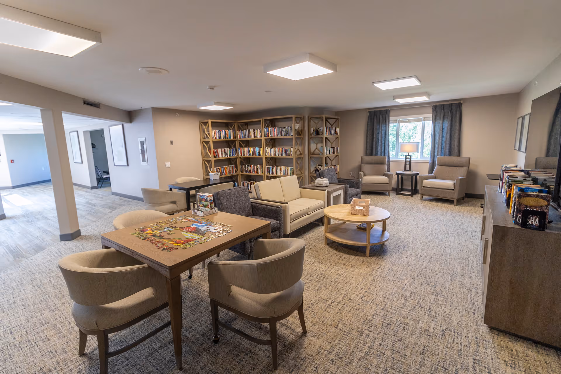 A bright and spacious common area in a senior living facility featuring a puzzle table with four chairs, a seating area with a loveseat and armchairs, a round coffee table, bookshelves filled with books, and a window with curtains letting in natural light.