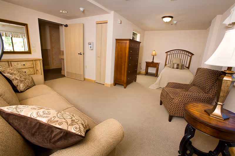 A cozy senior living bedroom with a single bed, wooden headboard, and beige bedding. The room includes a wooden dresser, a nightstand with a lamp, a patterned armchair, a round wooden side table with a lamp, and a beige sofa with decorative pillows. The walls are light-colored, and there is a mirror above a chest of drawers near the open door.