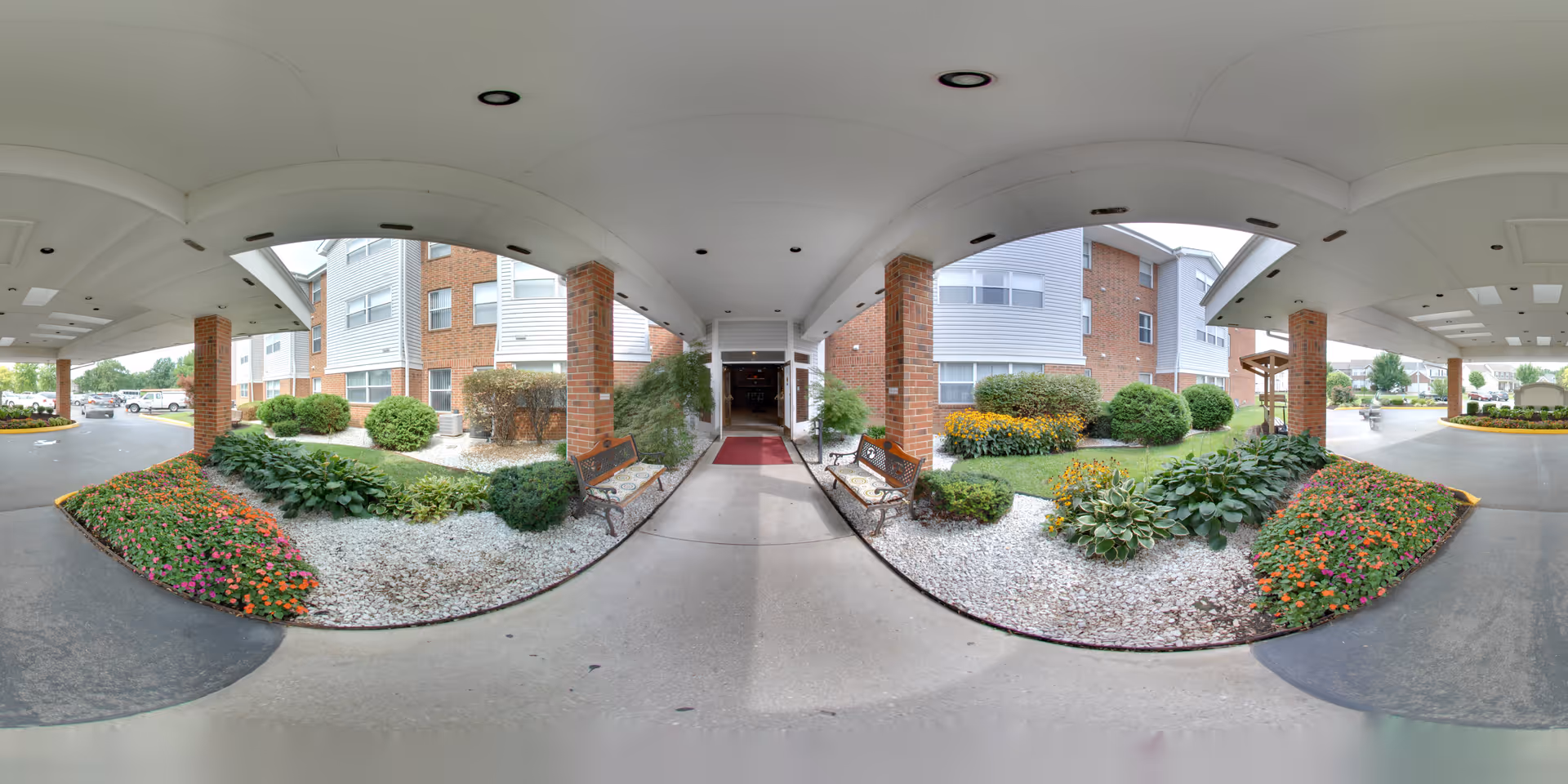Covered porte-cochere and entrance to a brick senior living building with landscaped flower beds, benches, and a walkway.
