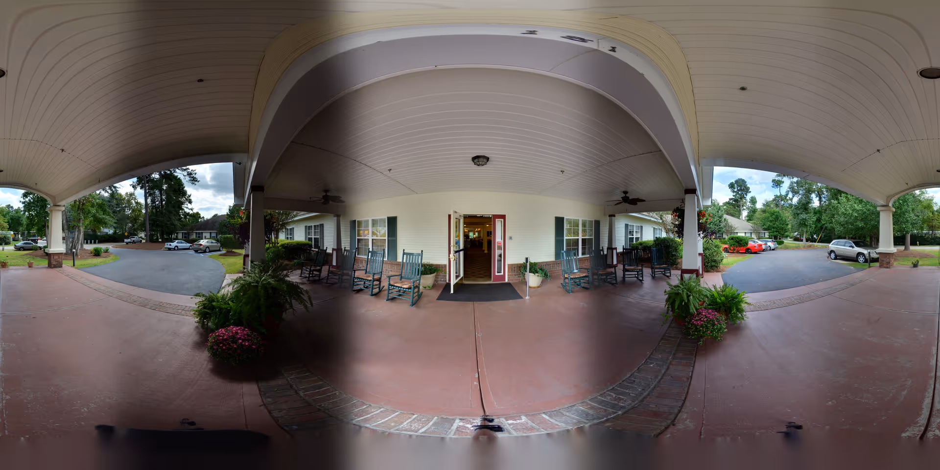Covered entrance area of a senior living facility with rocking chairs arranged on both sides near the entrance door. Potted plants and flowers decorate the space. The driveway and parking area with several cars are visible beyond the covered entrance. Trees and greenery surround the parking lot.