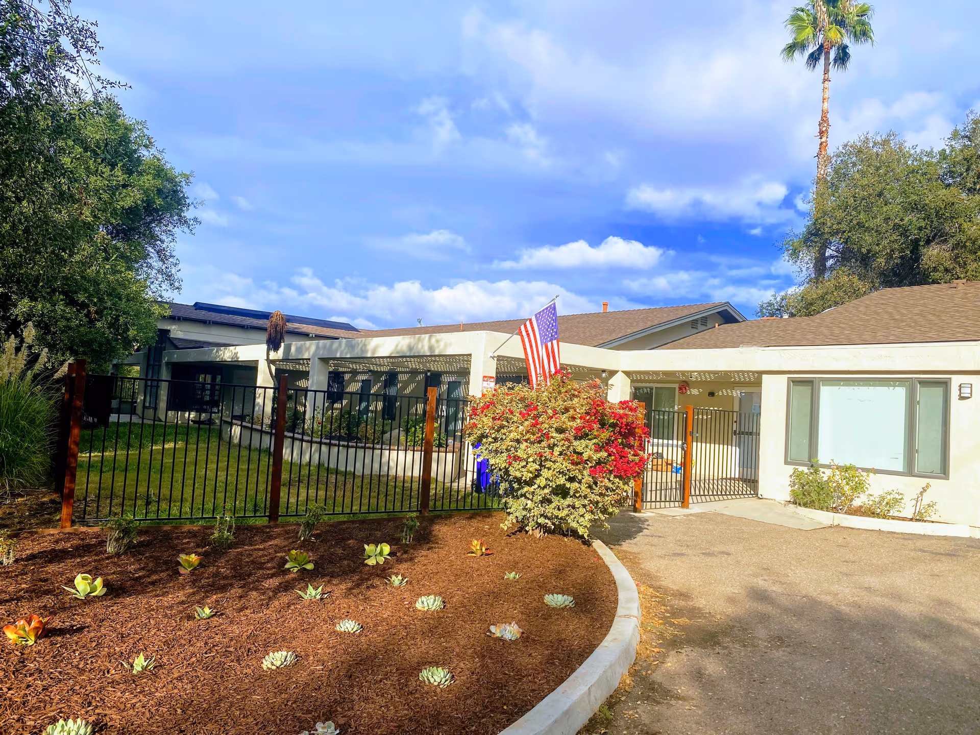 Exterior view of a single-story assisted living facility building with a fenced garden area, an American flag, various plants and shrubs, and a partly cloudy blue sky.
