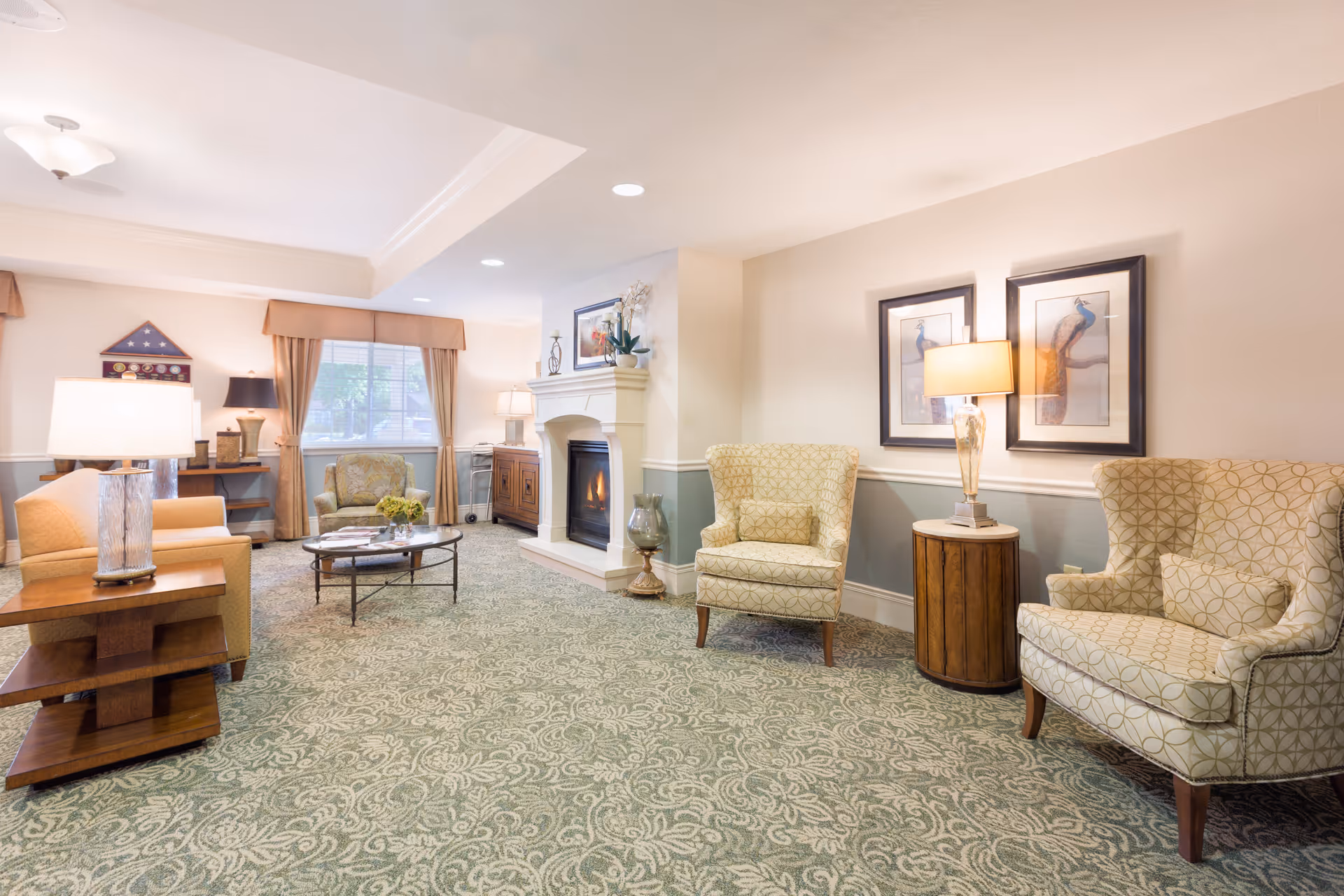 A cozy living room area in an assisted living facility featuring patterned carpet, two upholstered armchairs with matching cushions, a round wooden side table with a lamp, a fireplace with a mantel decorated with a plant and framed artwork, a coffee table with flowers, and a window with curtains allowing natural light.