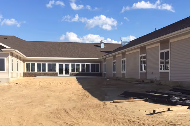 Exterior view of a single-story building under construction with beige siding, multiple windows, and a brown roof under a partly cloudy sky. The ground in front is bare dirt with some construction materials scattered around.