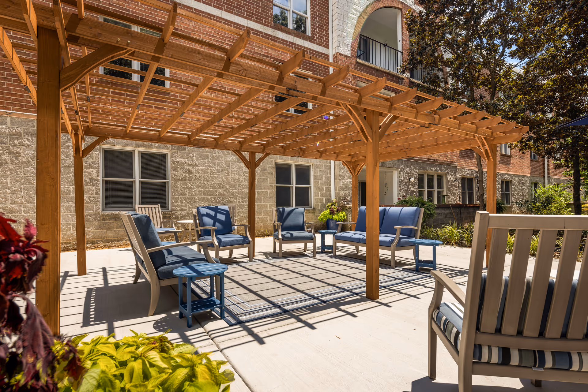 Outdoor patio area at Signal Mountain Senior Living featuring a wooden pergola with blue cushioned chairs and small tables arranged underneath. The patio is adjacent to a brick and stone building with windows, surrounded by some greenery and trees.