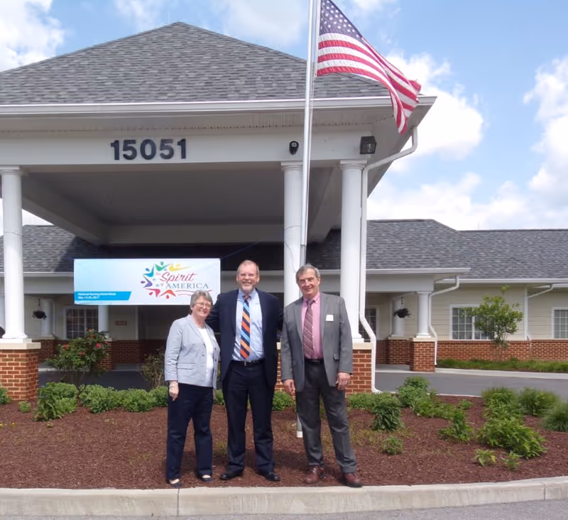 Three people stand under the covered entrance of Abingdon Health & Rehab Center (address 15051) beside an American flag and a 'Spirit of America' banner.