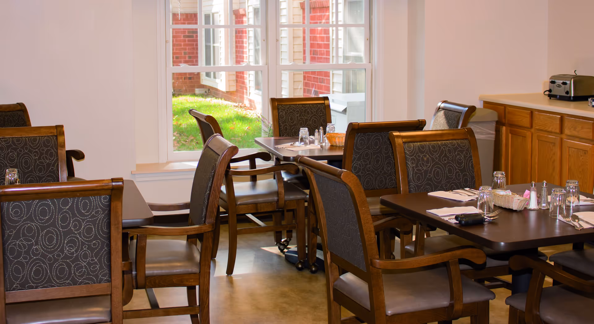 A dining area with several dark wood tables and chairs with patterned upholstery. The tables are set with glasses, napkins, and condiments. There is a large window showing a green lawn outside, and a wooden cabinet with a toaster on top is visible on the right side.