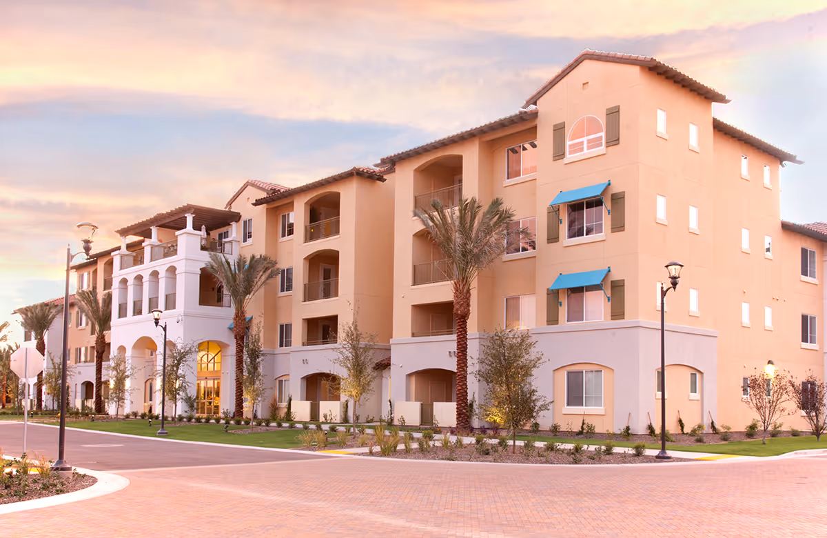 Exterior view of a multi-story senior living building with balconies, palm trees, and a brick-paved roadway.