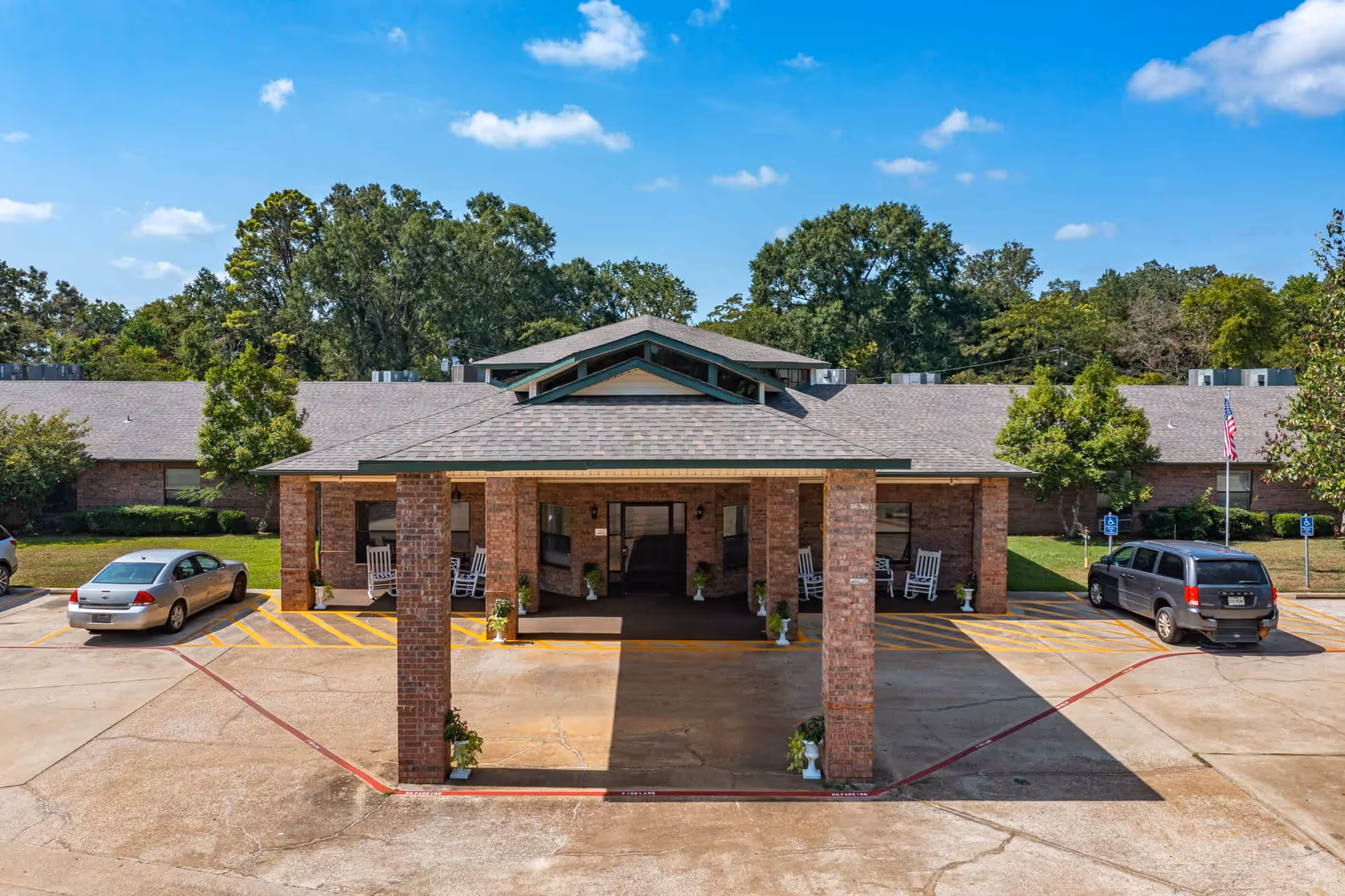 Front exterior view of Summit Nursing & Rehab of San Augustine showing a covered entrance with brick pillars, rocking chairs on the porch, parked cars, and trees in the background under a blue sky with some clouds.