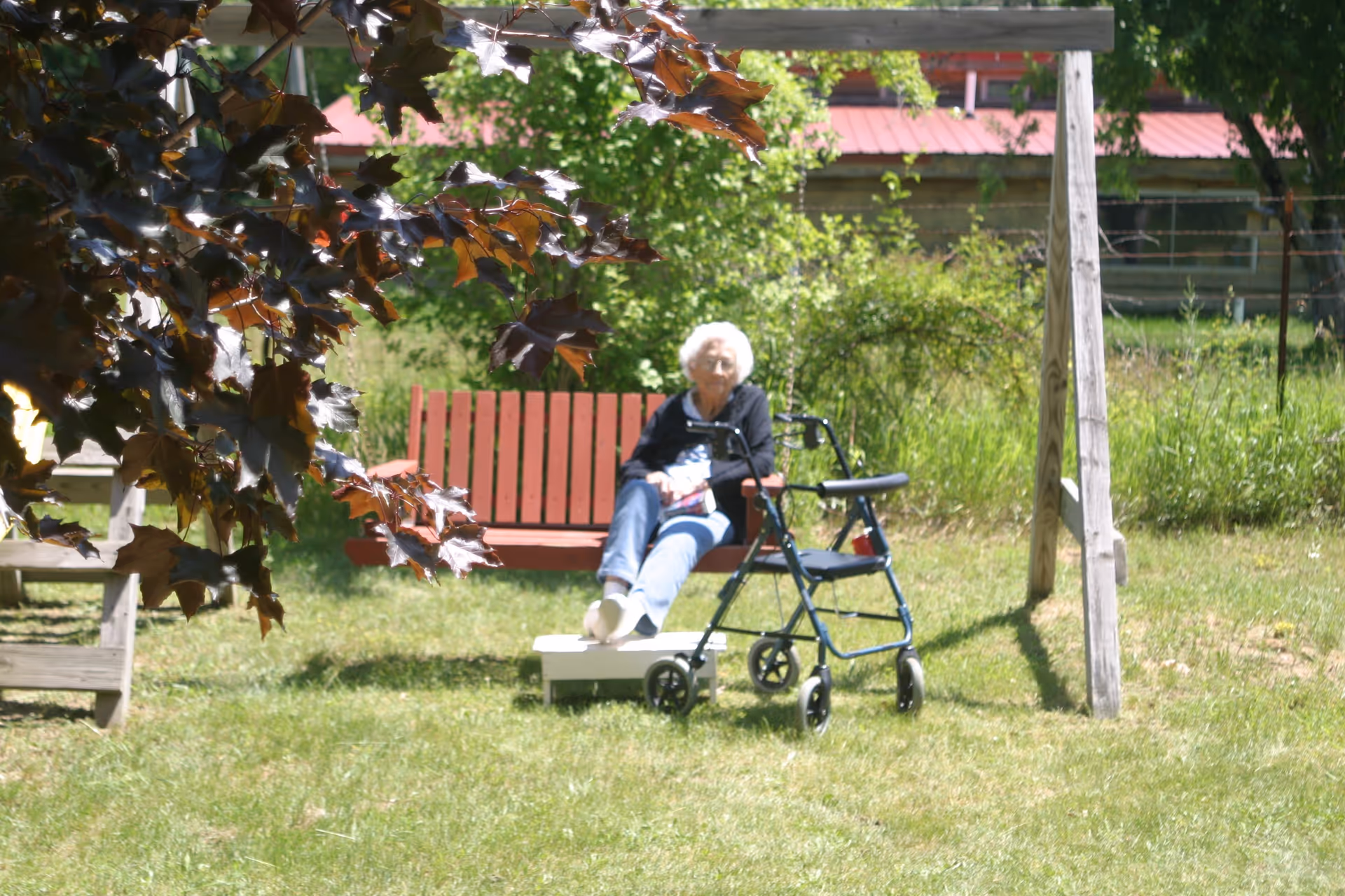 An elderly woman sitting on a red wooden swing in a grassy outdoor area with trees and bushes around. A blue walker is positioned in front of her. The background includes a wooden fence and a building with a red roof.