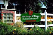 Green sign reading 'Heritage Village Retirement Campus' mounted on a white fence with greenery and flowers in front, and a brick structure with glass blocks to the left.