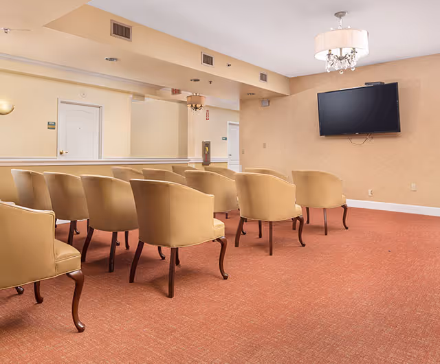 A small conference or meeting room with two rows of beige upholstered chairs facing a wall-mounted flat screen TV. The room has beige walls, a red carpet, and two ceiling light fixtures with lampshades and crystal accents.