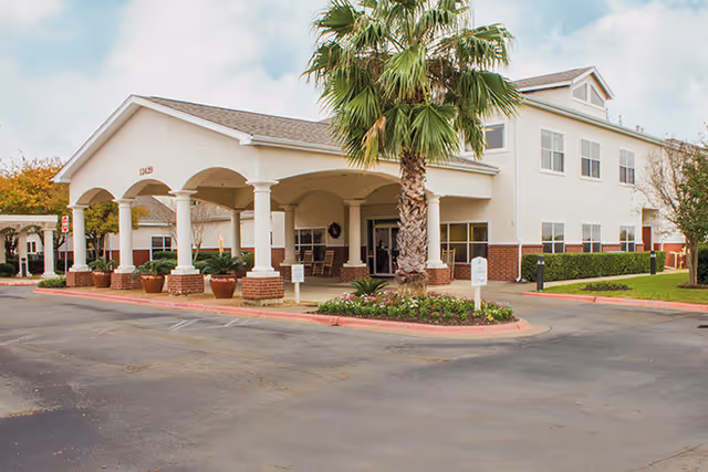 Front entrance of a two-story senior living building with a covered porte-cochère, palm tree, and landscaped driveway.
