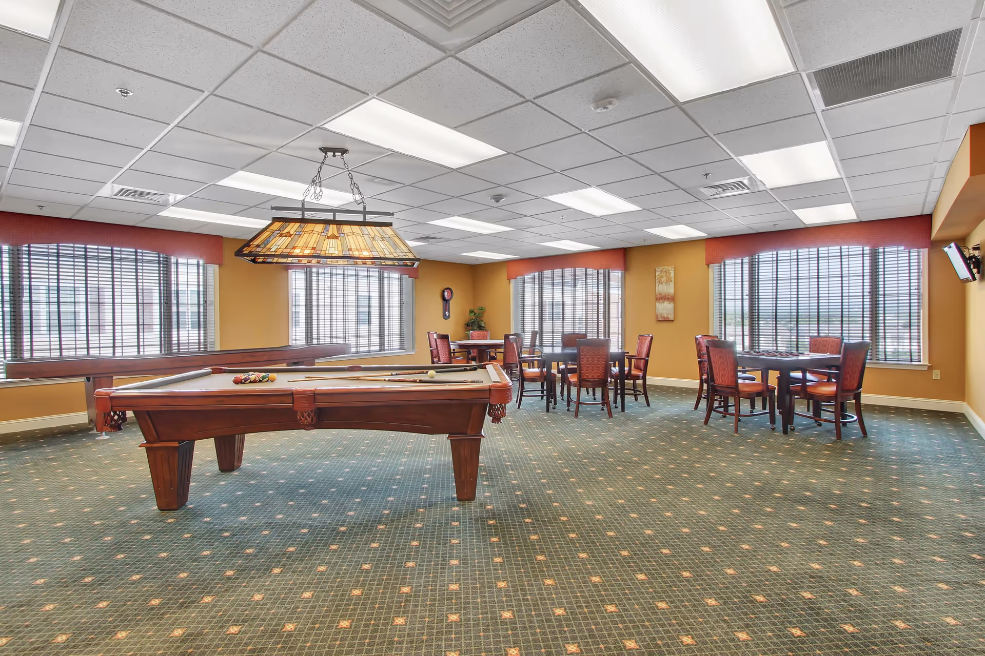 A spacious recreational room with a pool table in the center, a shuffleboard table along the left wall, and several tables with chairs near large windows with blinds. The room has a green patterned carpet, yellow walls, and a ceiling with fluorescent lights and a hanging lamp above the pool table.