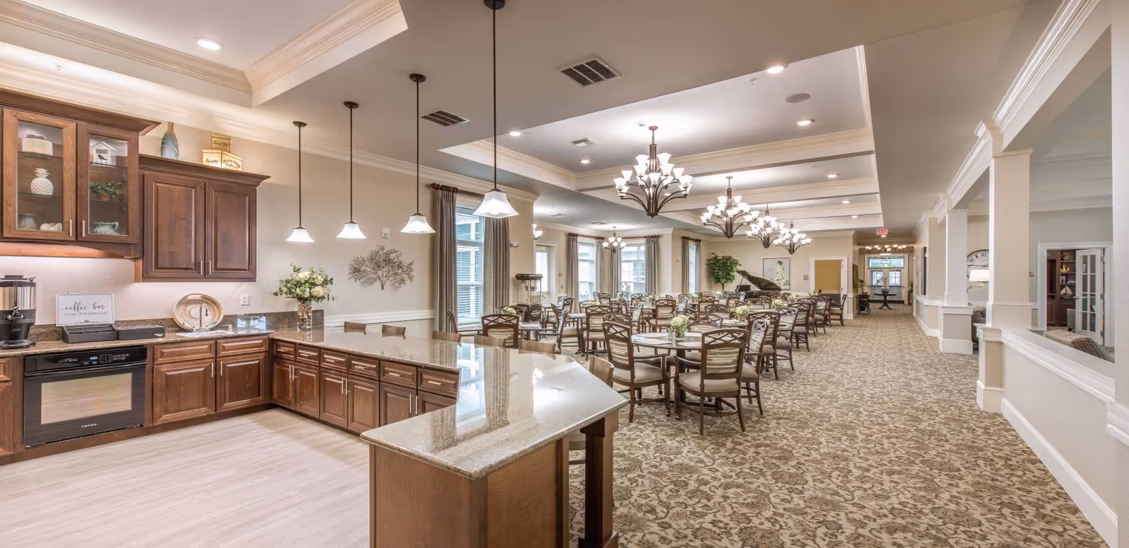 Open dining room with multiple tables and chairs adjacent to a serving kitchen and island under chandeliers.